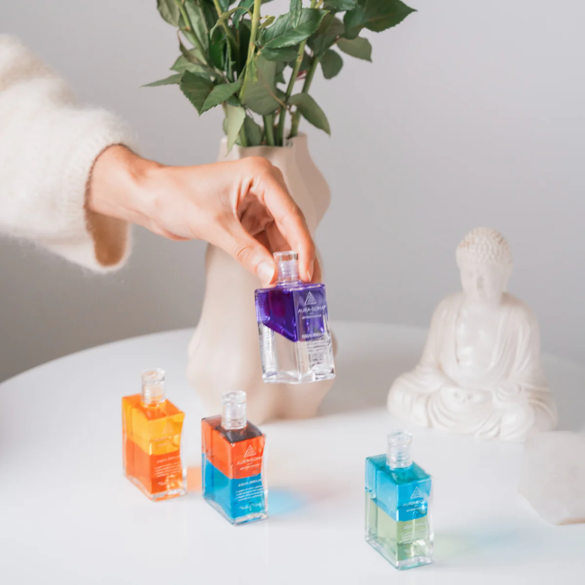 A hand selecting an Aura-Soma Equilibrium bottle from a table with other bottles and a Buddha statue.